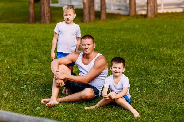 Жорстокий чоловік і діти в білих футболках сидять на зеленій траві влітку — стокове фото