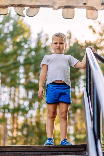 Sju år gammal pojke i full höjd i en vit t-shirt och blå shorts på en suddig naturlig bakgrund tittar på kameran på sommaren — Stockfoto