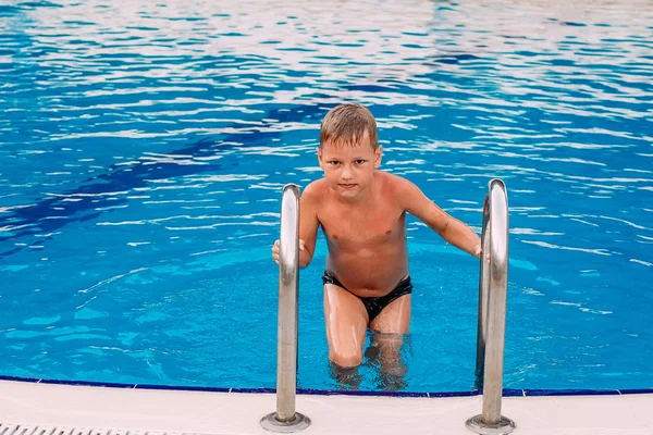 Загорелый мальчик вылезает из открытого синего бассейна и смотрит в камеру — стоковое фото