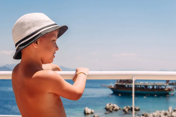 Un niño con un sombrero blanco sosteniendo la barandilla en el barco y mirando al mar . —  Fotos de Stock