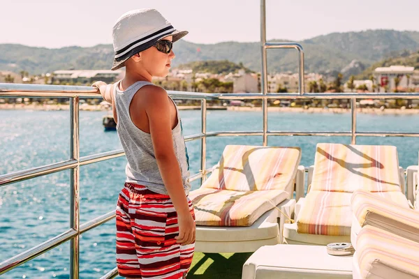 En pojke i en vit hatt och shorts som håller räcket på skeppet och tittar på havet. — Stockfoto