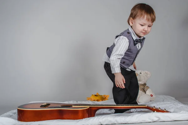 Two year old Caucasian boy in bow tie and vest with acoustic guitar on grey background — Stock Photo, Image