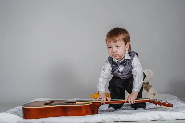 Милый белый мальчик в бабочке и жилете с гитарой на сером фоне — стоковое фото