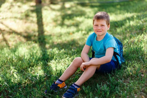 Счастливый улыбающийся школьник с рюкзаком сидит на зеленой траве — стоковое фото