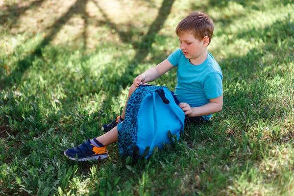 Feliz estudiante de ocho años de edad, está sentado en la hierba verde y abrió su mochila —  Fotos de Stock