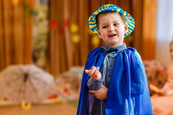 青いスーツを着た陽気な5歳の子供と、幼稚園のぼやけた背景に踊る帽子 — ストック写真