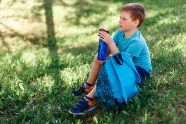 Çocuk elinde bir şişe suyla çimenlerin üzerinde oturuyor.