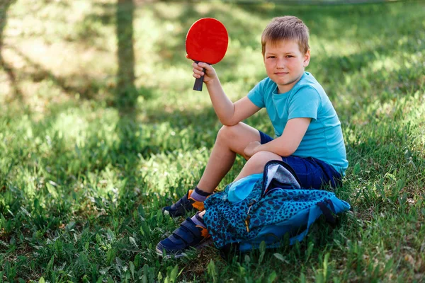 Восьмилетний мальчик сидит на зеленой траве и держит ракетку для настольного тенниса в руках — стоковое фото