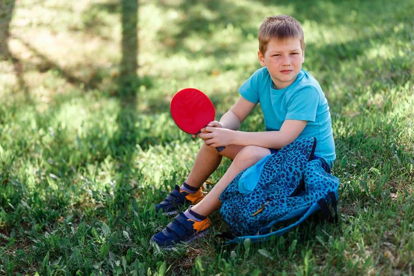 8-летний спортивный мальчик в кроссовках сидит на зеленой траве и держит ракетку для настольного тенниса в руках — стоковое фото