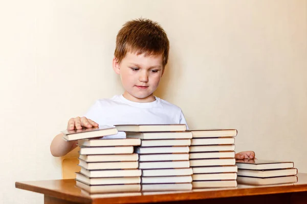 Un ragazzo diligente siede a una scrivania e mette libri su pile . — Foto Stock