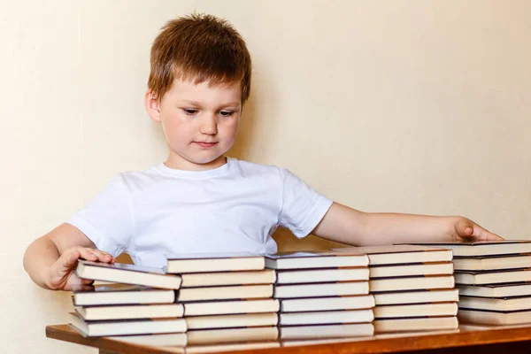 Un simpatico bambino di sei anni si siede a una scrivania e mette libri su pile . — Foto Stock