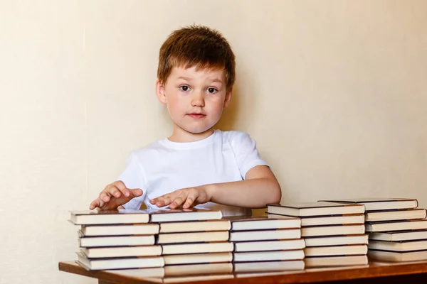 Carino ragazzo di sei anni seduto a una scrivania con pile di libri . — Foto Stock