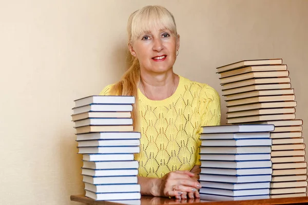 Усміхнена п'ятдесятирічна жінка в жовтому светрі сидить за столом з книгами — стокове фото