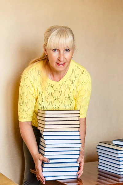 smiling fifty-year-old woman librarian in a yellow jacket raises a large stack of identical books
