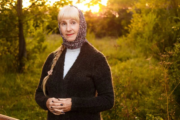 Улыбающаяся пятидесятилетняя женщина в шарфе на голове и кардигане, смотрящая на камеру летом на закате — стоковое фото