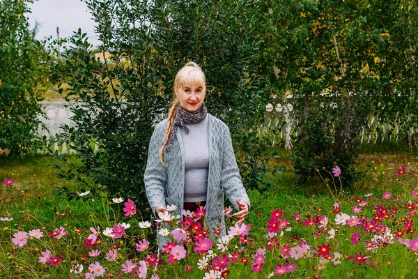 Пятидесятилетняя улыбающаяся женщина в сером кардигане стоит среди цветущих красочных астеров в саду — стоковое фото