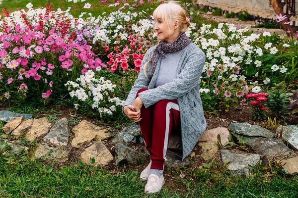 Пятидесятилетняя привлекательная блондинка в сером кардигане сидит среди цветущих цветущих петуний — стоковое фото