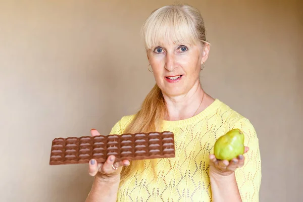 Емоційна літня кавказька жінка тримає шоколад і фрукти, жінка на дієті — стокове фото