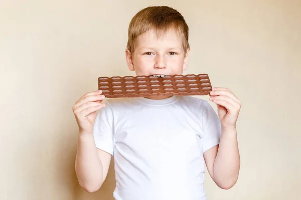 Семилетний белый мальчик в белой футболке и огромном молочном шоколаде. дети и сладости — стоковое фото