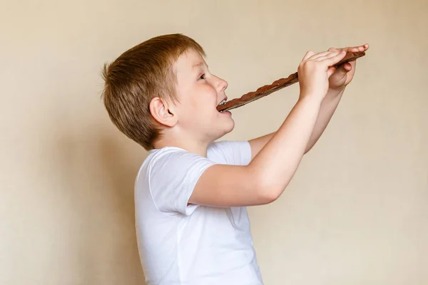 Sedmiletý kavkazské dítě v bílém tričku kousne obrovskou mléčnou čokoládu — Stock fotografie