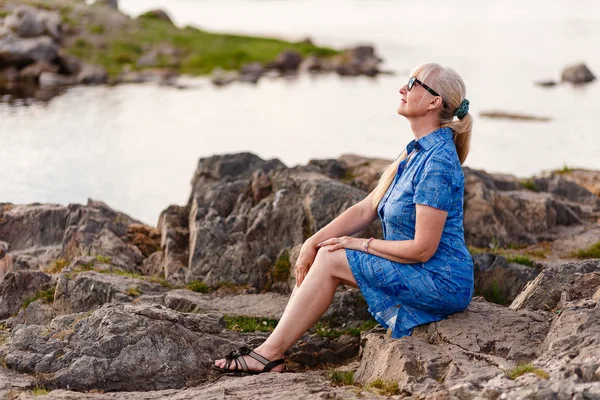 Vacker kvinna pensionsåldern i profil i en blå klänning sitter på en sten nära floden vid solnedgången — Stockfoto