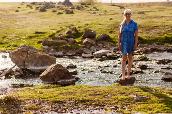 Усміхнена красива жінка пенсійного віку в блакитній сукні, що стоїть на камені біля річки на заході сонця — стокове фото