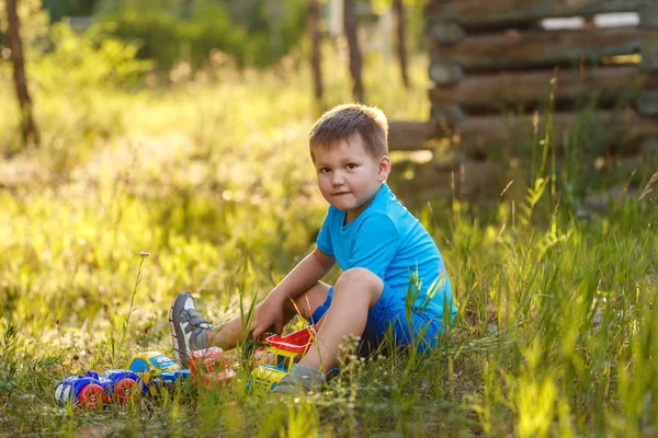 cute five-year-old boy in blue clothes playing with cars in the summer sitting on the grass