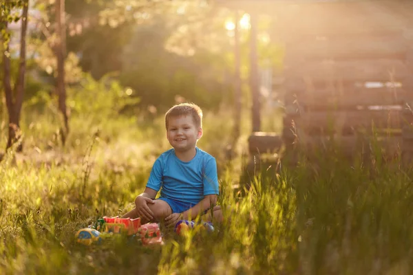 Leende femårig pojke i blå kläder leker med bilar på sommaren sitter på gräset i ljuset — Stockfoto