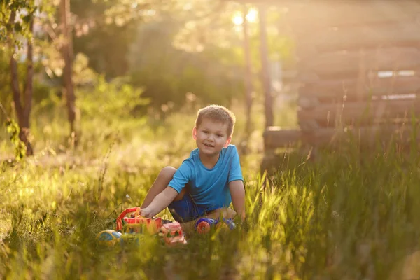 Söt femårig pojke i en blå t-shirt och shorts leker med plast bilar på sommaren sitter på gräset i parken i det varma ljuset — Stockfoto