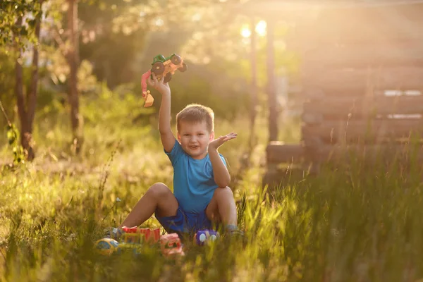 Весёлый эмоциональный пятилетний мальчик в синей футболке и шортах, играющий с пластиковыми игрушками летом, сидя на траве в парке на закате солнца — стоковое фото