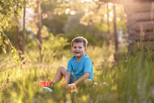 Кавказский пятилетний мальчик в голубой одежде летом сидит на траве и играет с игрушками в саду на солнце — стоковое фото