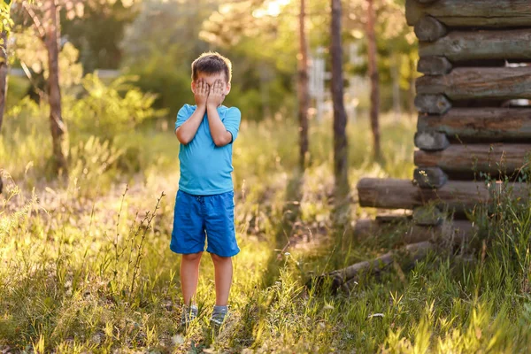 Пятилетний мальчик в голубой одежде закрывает лицо руками летом при свете заката — стоковое фото
