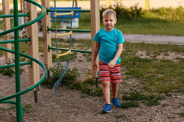 Семилетний мальчик в бирюзовой футболке и красных шортах летом на детской площадке — стоковое фото