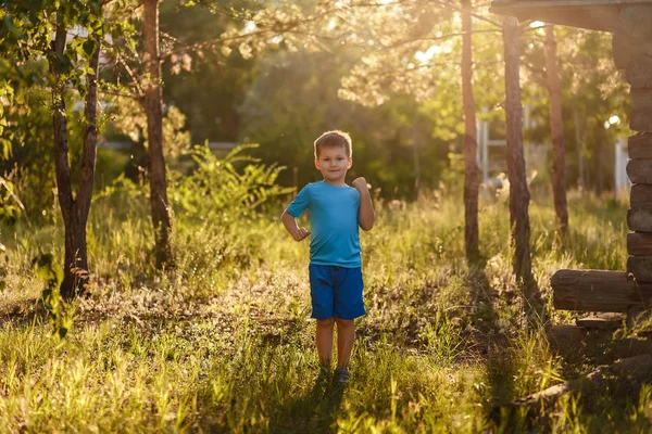 Sportowy pięcioletni kaukaski chłopiec w niebieskim t-shirt i szorty stoi w parku w tylnej światło słoneczne w lecie — Zdjęcie stockowe
