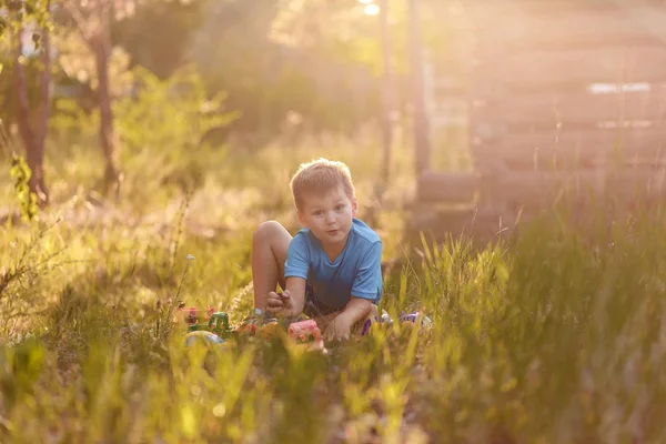 Весёлый эмоциональный пятилетний мальчик в голубой футболке и шортах, сидящий на траве летом и играющий с игрушками — стоковое фото