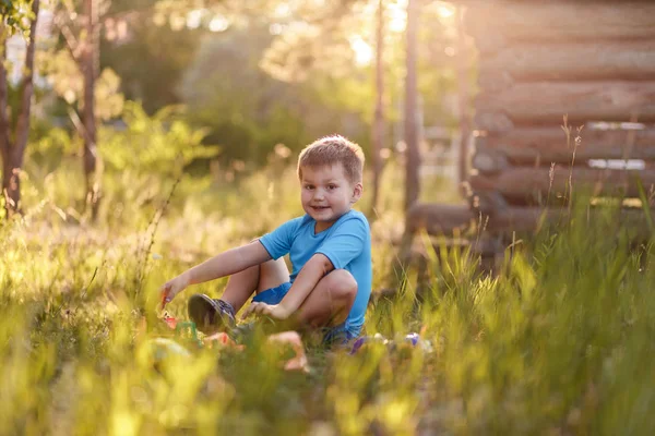 Милый пятилетний мальчик в голубой футболке и шортах, сидящий на траве летом и играющий с игрушками в саду на солнце — стоковое фото