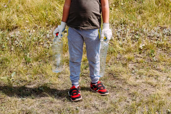 Мальчик в рабочих перчатках удаляет пластиковые обломки, держа в руках пустые пластиковые бутылки. экологические проблемы — стоковое фото