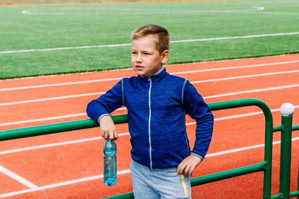 Восьмилетний мальчик в спортивном костюме с бутылкой воды в руках стоит на спортивном стадионе — стоковое фото