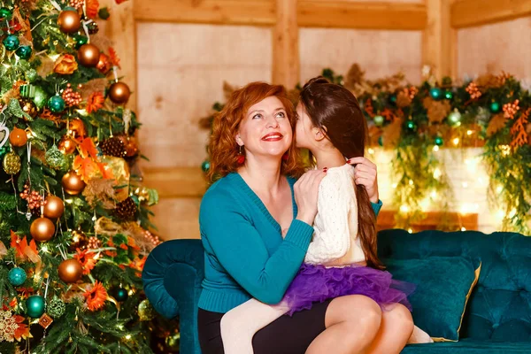 En flicka med mörkt hår sitter i sin mors knä och viskar en hemlighet i hennes öra i julklapp — Stockfoto