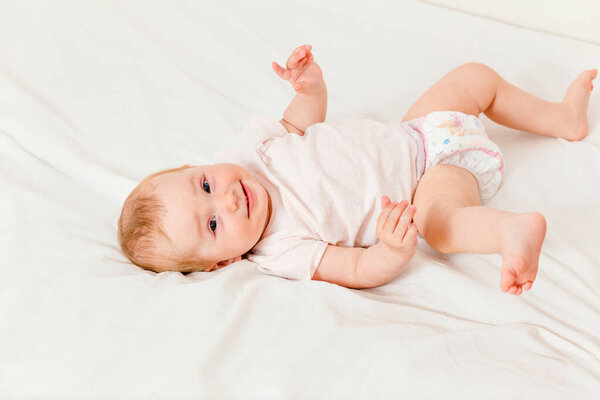 Cute little baby girl in a diaper and T-shirt lies on a white sheet on the bed