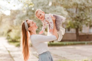 Uzun saçlı genç bir kadın açık havada gür bir yaz elbisesi içinde gülümseyen küçük bir kız çocuğu yetiştiriyor.