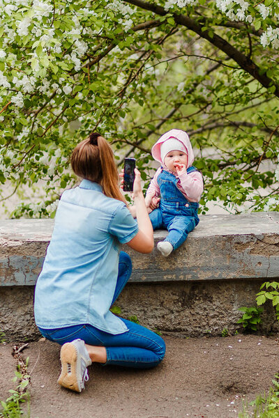 Мама фотографирует маленькую дочь по телефону на улице
