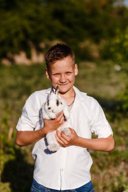 Gülümseyen 10 yaşındaki çocuk elinde küçük beyaz bir tavşan tutuyor.