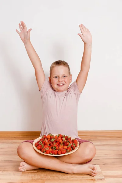 a happy boy sits on the floor with a large plate of strawberries and raises his hands in the air