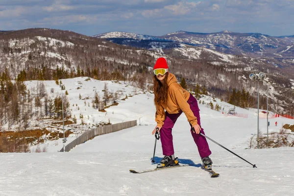 戴着红帽子和眼镜的年轻漂亮的女人站在山上滑雪板上 — 图库照片