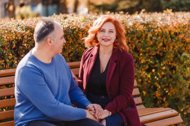 Gülümseyen orta yaşlı bir kadın ve bir erkek sonbaharda bir bankta otururken el ele tutuşuyorlar. Çift