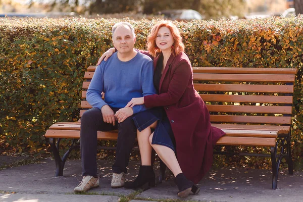 一位身穿蓝色毛衣的50岁男子和一位身穿勃艮第外套的女士坐在公园的长椅上 看着摄像机 — 图库照片