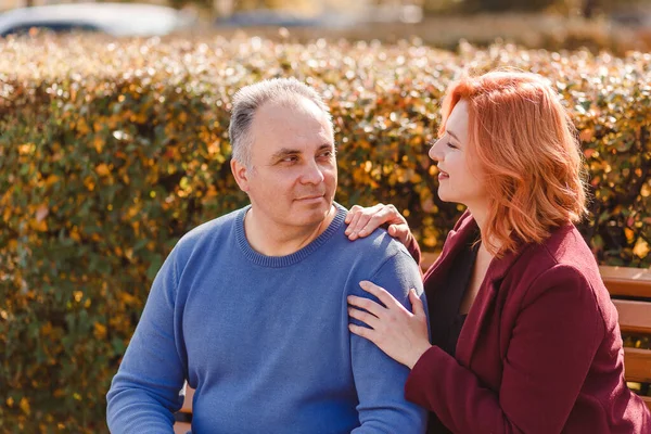 A 50-year-old man in a blue sweater and a woman in a burgundy coat sit on a park bench in the close