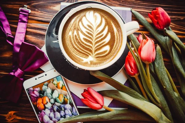Kahverengi arka planda bir fincan kahve, telefon ve çiçekler.