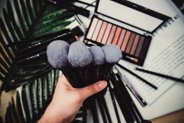 Makeup brushes, palette of shadows and green leaves on a white wooden background